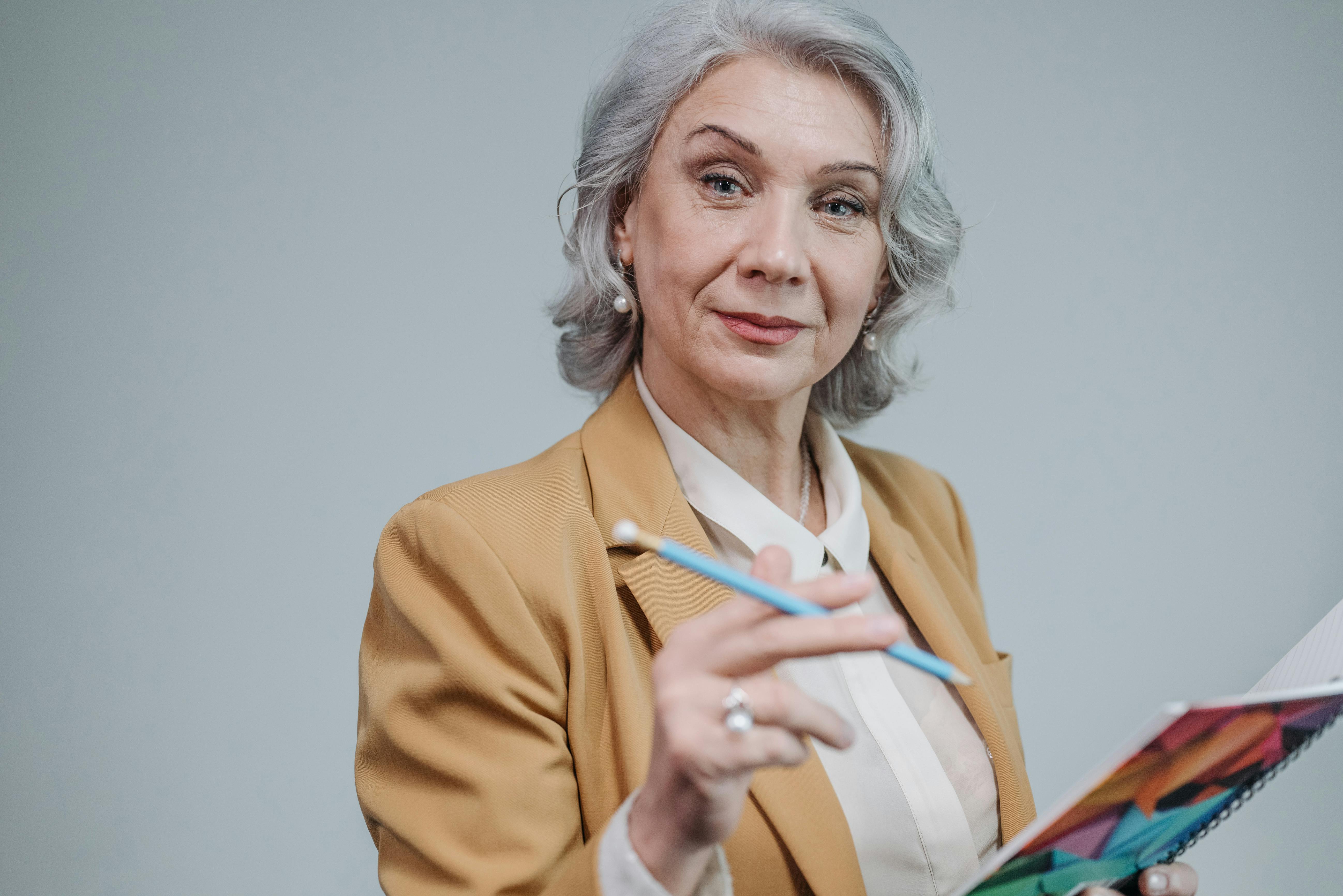 Portrait of a senior woman in a brown blazer holding a notebook and smiling warmly.